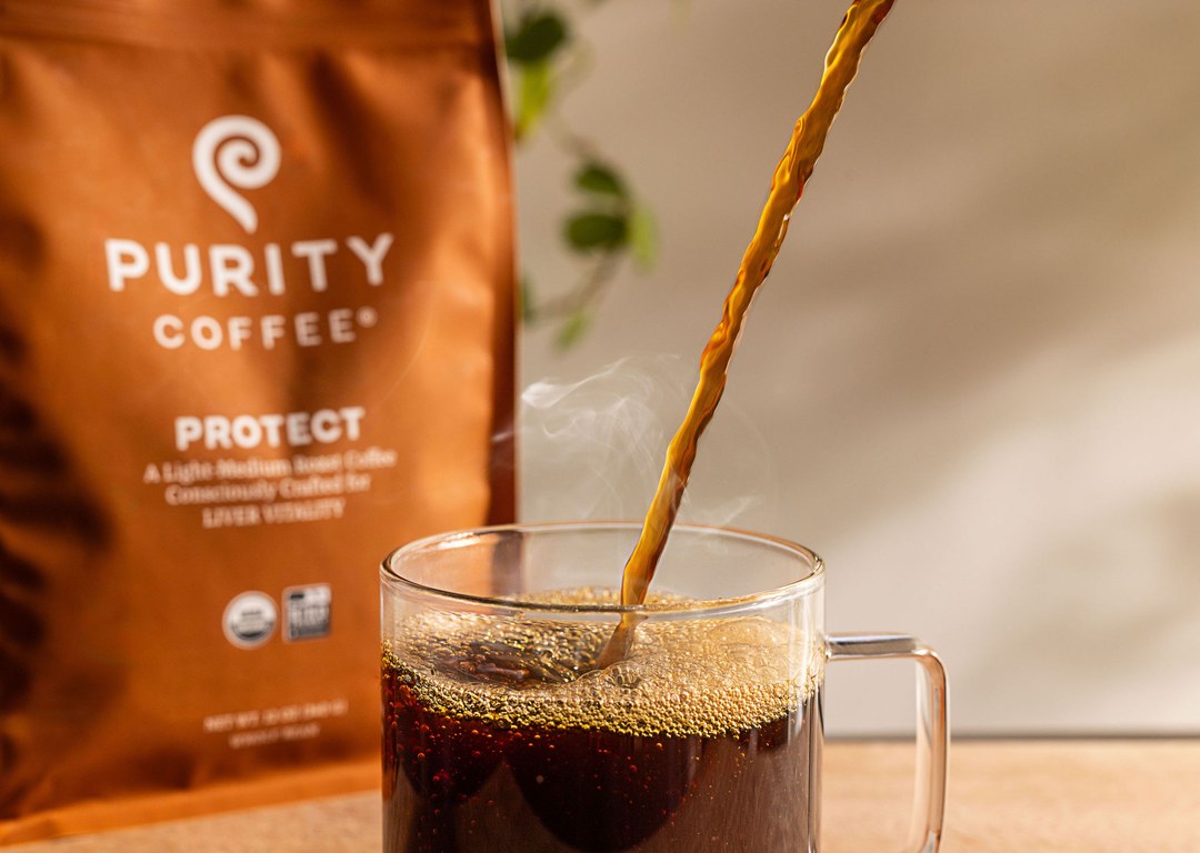 Hot coffee pouring into a glass mug beside a bag of Purity Coffee PROTECT